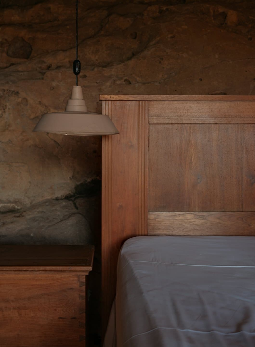Wooden bed with grey sheets sits against a stone wall.